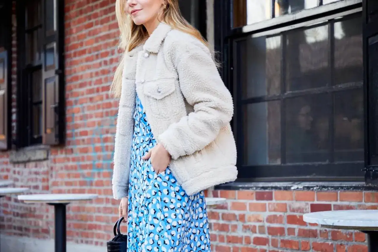 Une femme porte une robe courte bleue à motifs blancs et une veste en sherpa crème. Une idée parfaite pour savoir quelle veste porter avec une robe courte.