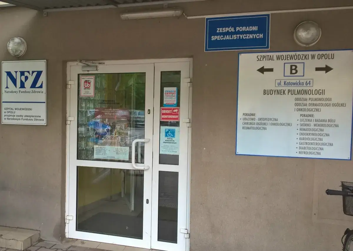Drzwi wejściowe do budynku szpitala z tablicami informacyjnymi NFZ i Szpitala Wojewódzkiego w Opolu.