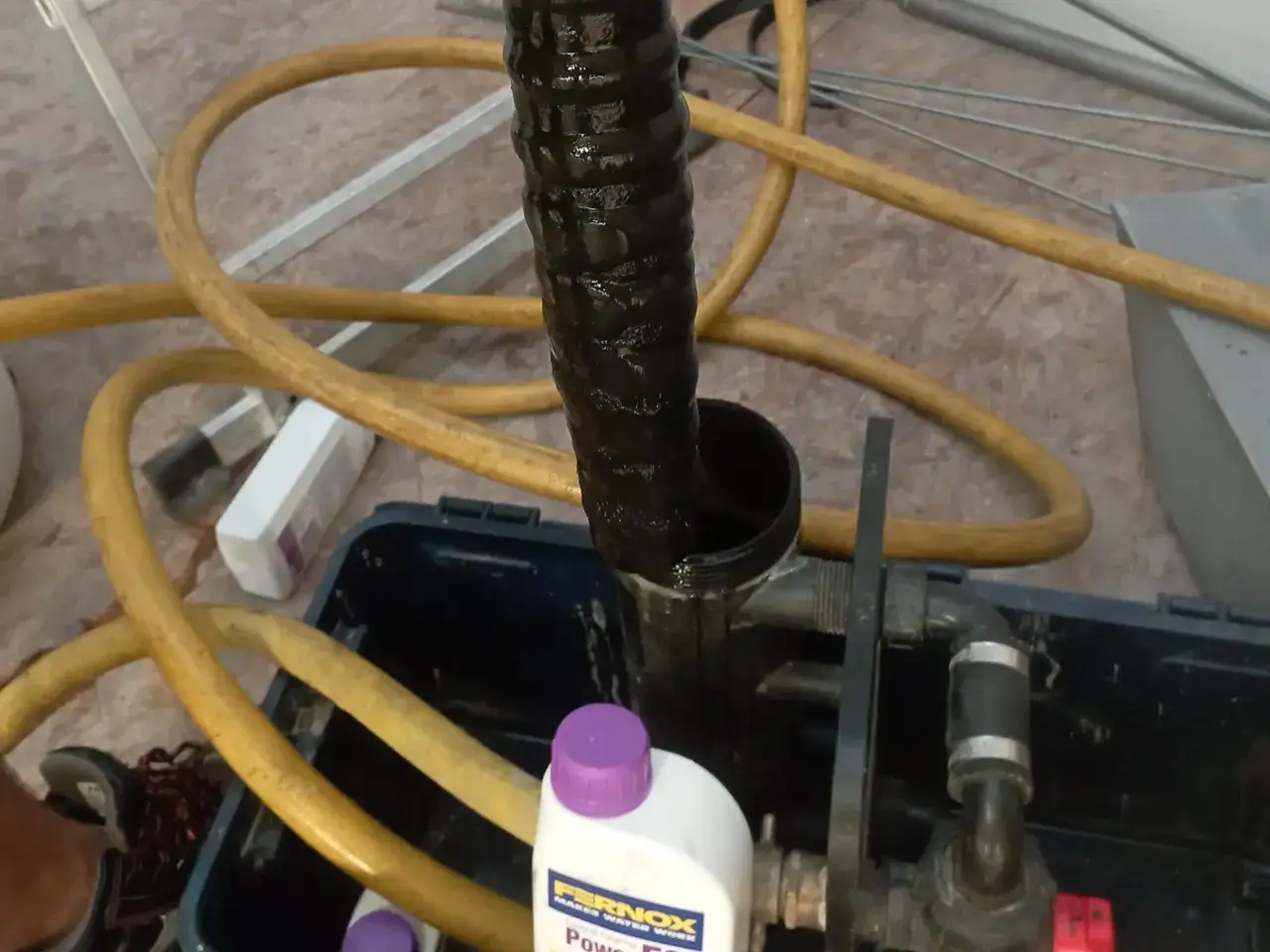 Płukanie instalacji grzewczej z użyciem preparatu Fernox. Widać wąż, pojemnik i butelkę z płynem. Cennik usług dostępny online.