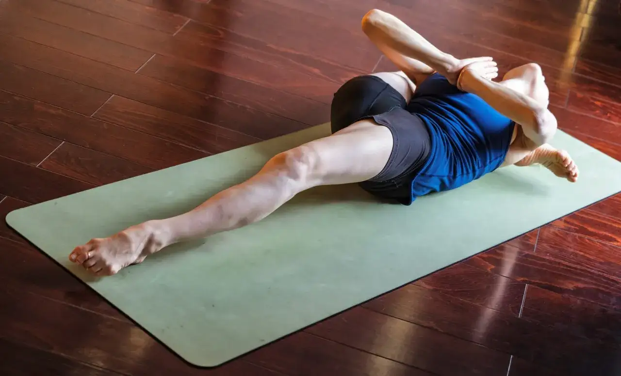 Pratique de l'ashtanga yoga : une personne en posture de torsion sur un tapis vert, jambes allong&eacute;es et bras crois&eacute;s.