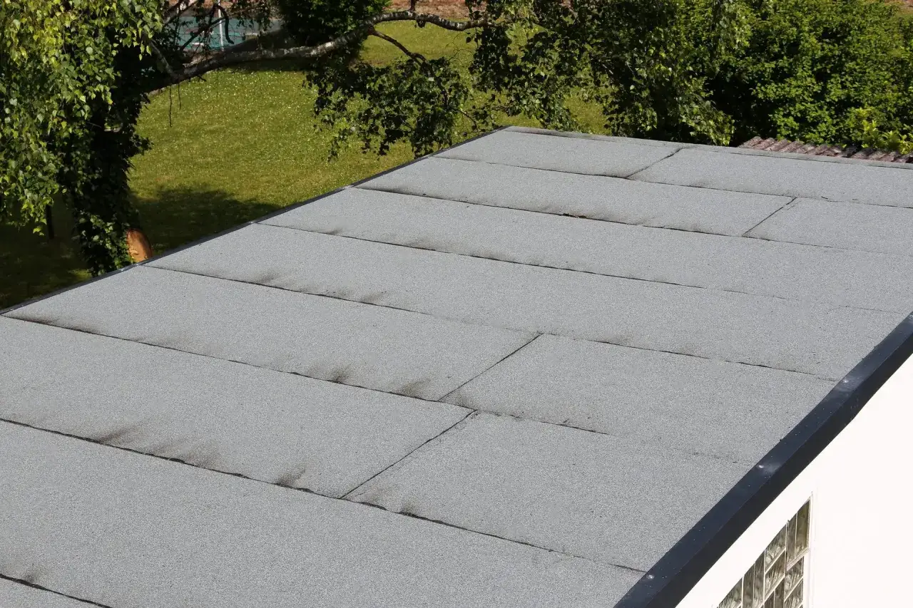 Beton garagendach abdichten: graue Bitumenbahnen auf einem Flachdach, daneben grüner Rasen und Bäume.