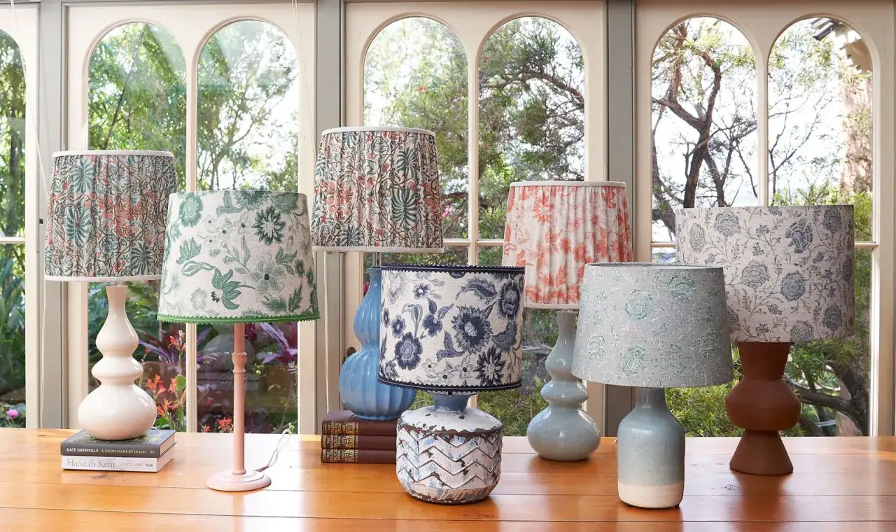 Plusieurs lampes avec des abat-jour en tissu aux motifs floraux et végétaux, dans des tons verts, bleus et corail, sur un bureau en bois devant une fenêtre.