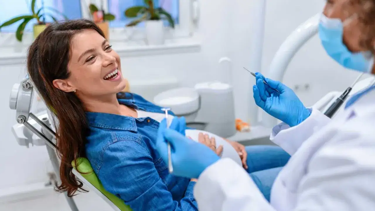 Uśmiechnięta ciężarna kobieta na fotelu dentystycznym, przygotowująca się do piaskowania zębów w ciąży.