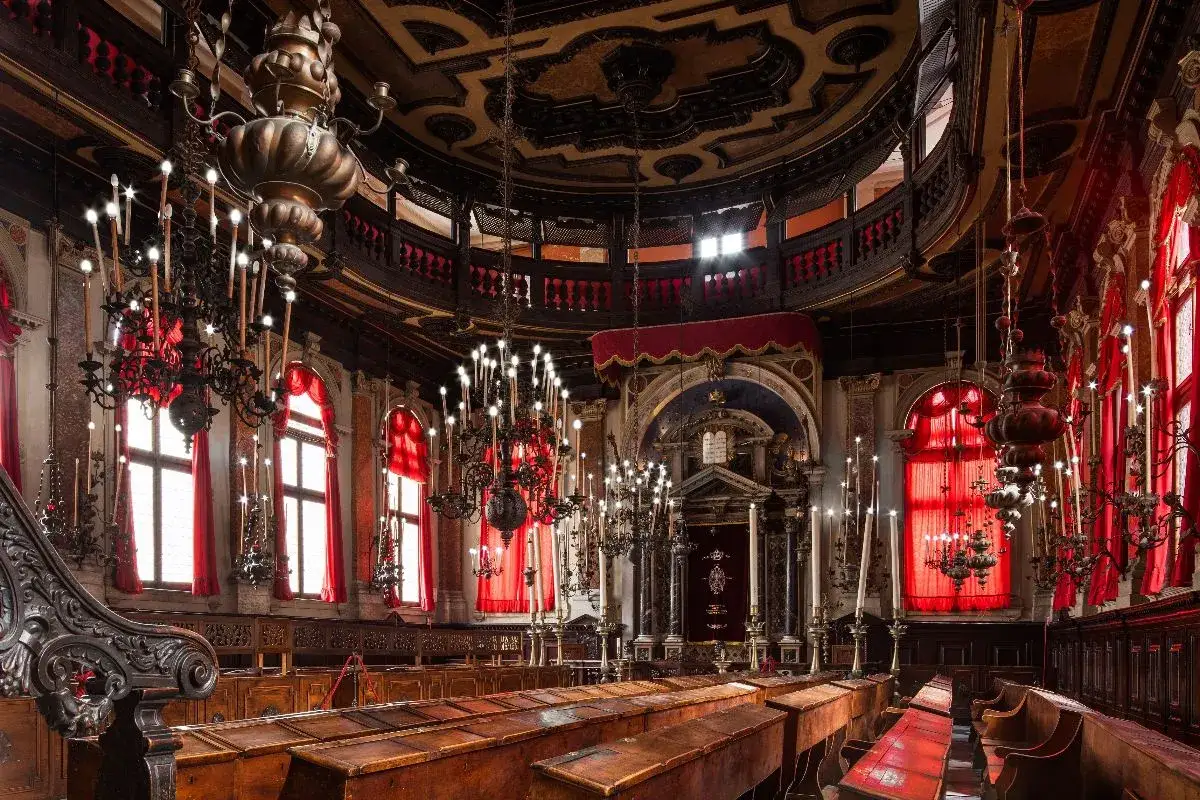 Interno sontuoso della sinagoga di Venezia, con panche in legno, candelabri elaborati e tende rosse che filtrano la luce.
