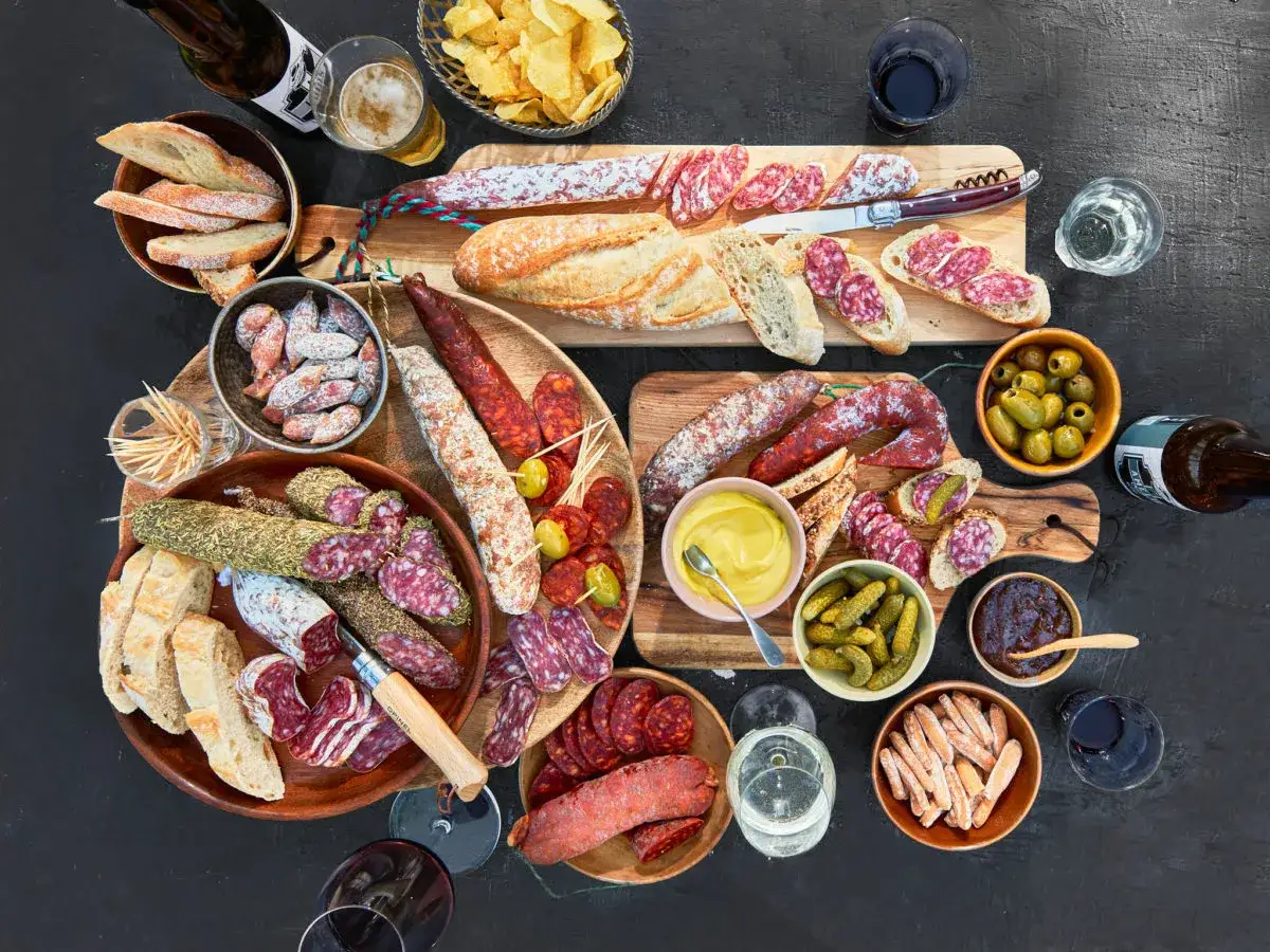 Eine reichhaltige **Wurstplatte anrichten** mit Baguette, Oliven, Cornichons, Senf und Bier.