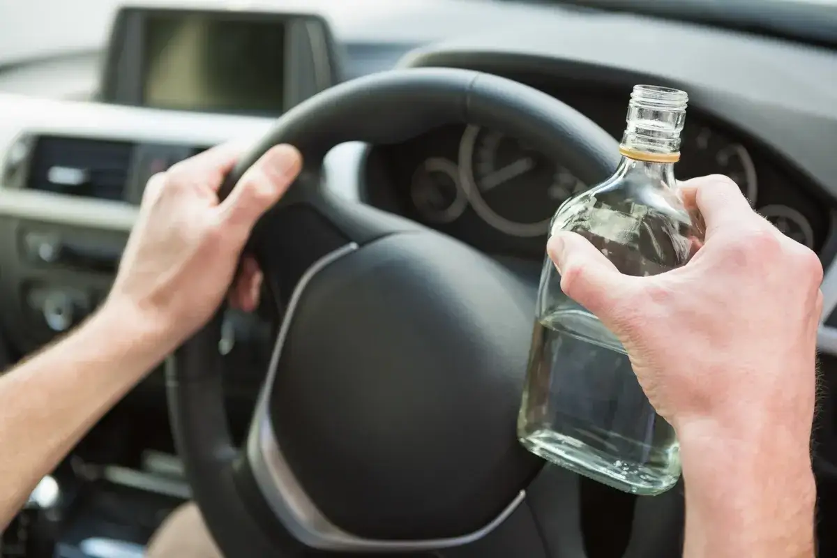 Ręce kierowcy trzymają kierownicę i butelkę alkoholu. Zastanawiasz się, ile się trzeźwieje po butelce wina? Nie ryzykuj.