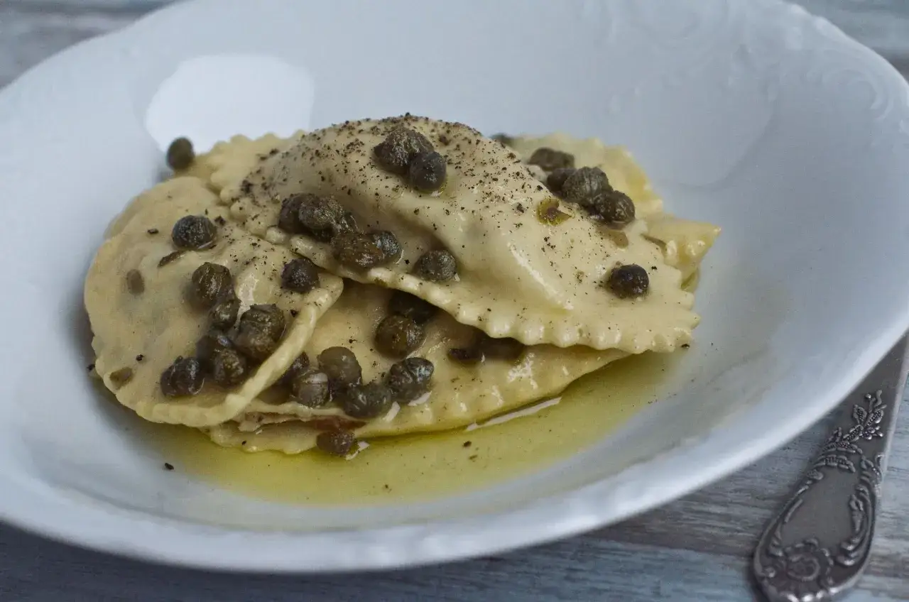 Pyszne ravioli z kurczakiem, polane oliwą i posypane kaparami i pieprzem, na białym talerzu.
