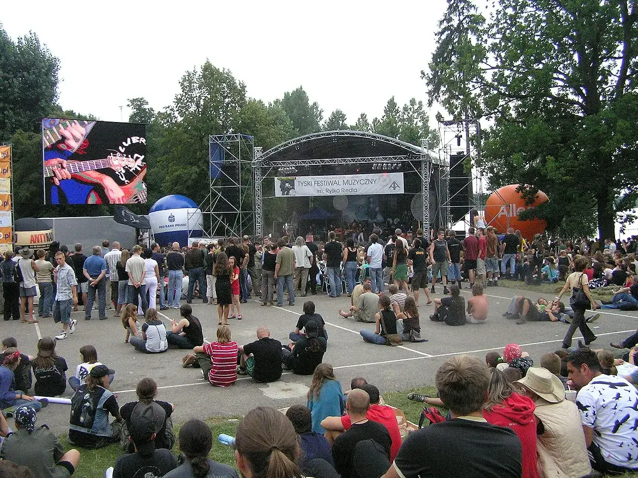Tyski festiwal muzyczny gromadzi tłumy fan&oacute;w. Na scenie gra zesp&oacute;ł, a na wielkim ekranie widać gitarzystę. Wszyscy cieszą się atmosferą festiwali muzycznych.