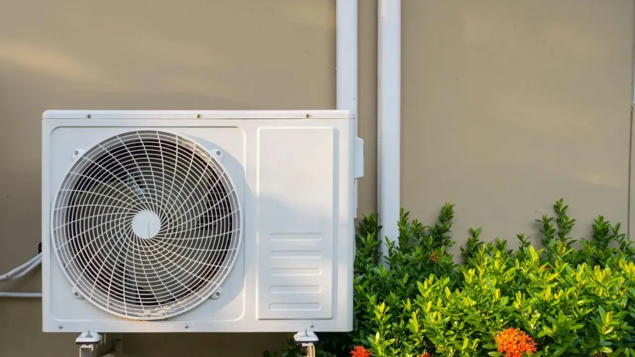 Außenunit einer Split-Klimaanlage an einer Wand, umgeben von grünen Pflanzen. Regelmäßige Wartung der Split-Klimaanlage sorgt für optimale Leistung.
