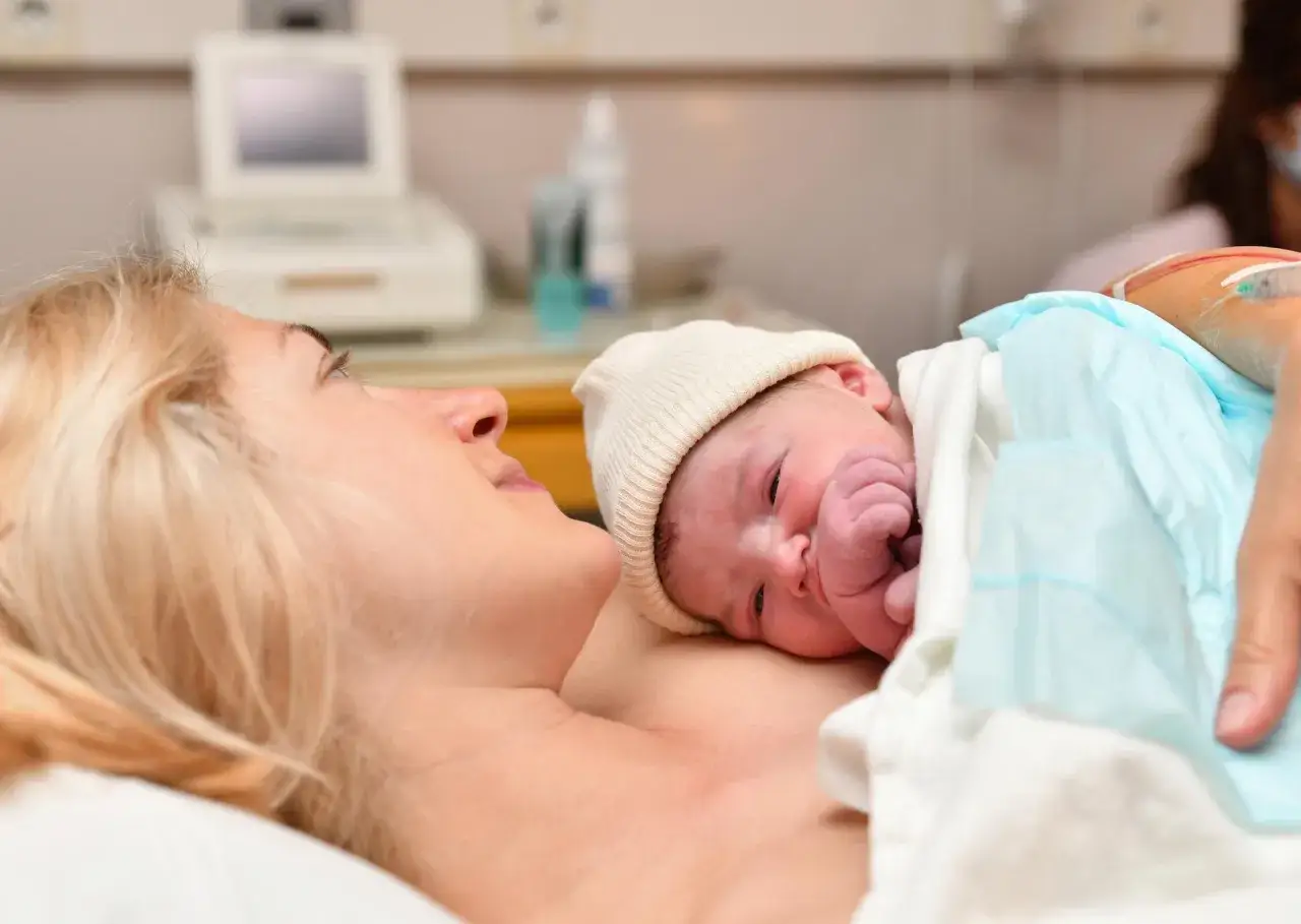 Nowa mama z noworodkiem na piersi. W tle aparat USG, jakby szukała odpowiedzi na pytania do położnej przed porodem.