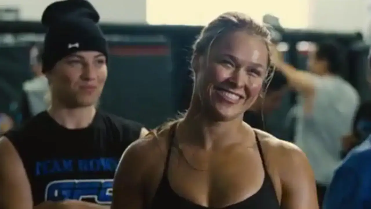 Ronda Rousey sorride in una scena di un film, con un uomo con un berretto nero sullo sfondo.