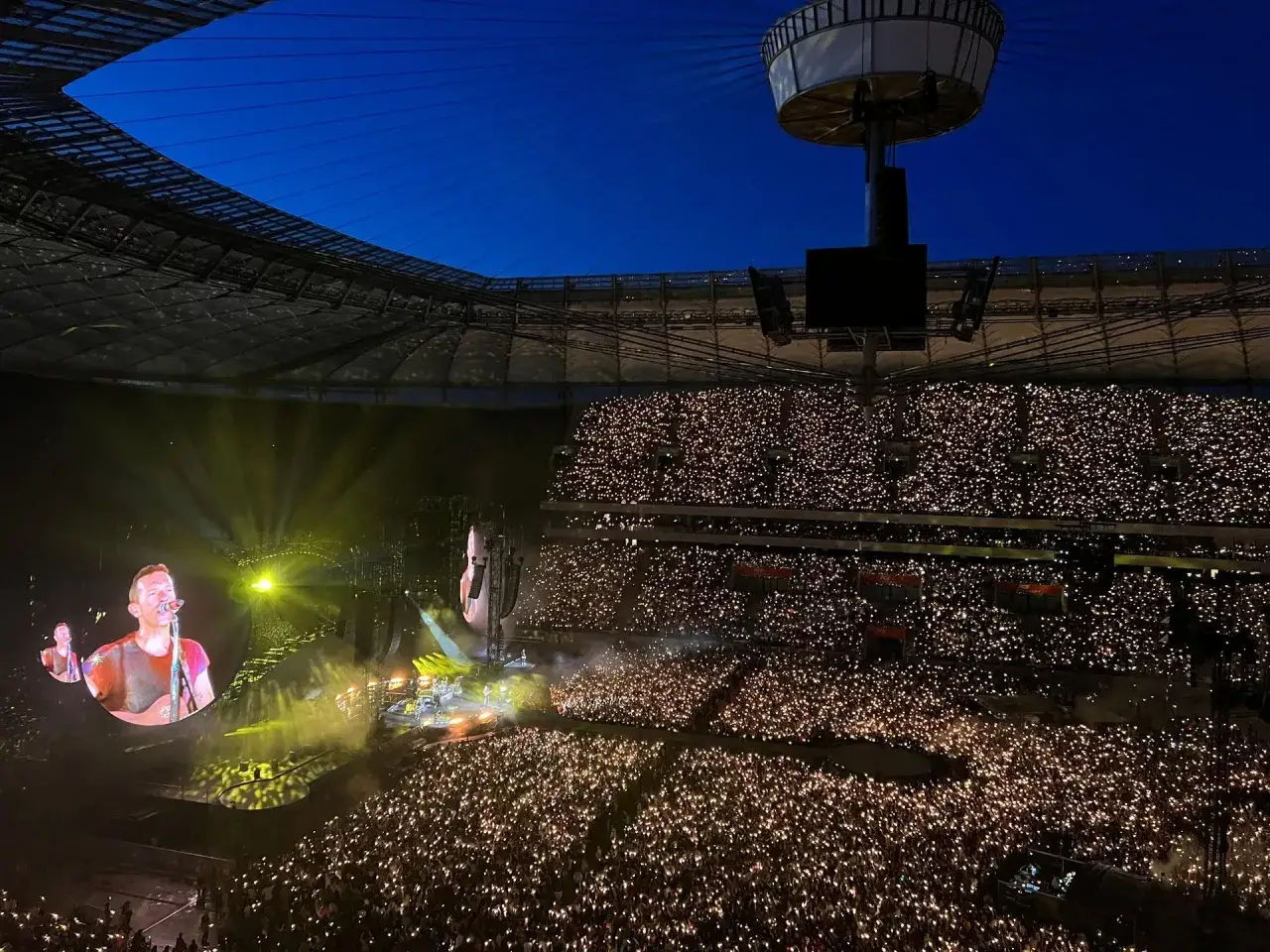 Koncert Coldplay w Polsce. Stadion wypełniony po brzegi, tysiące świateł w rękach fanów tworzą magiczną atmosferę. Na telebimie Chris Martin.