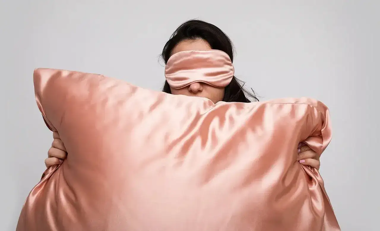 A person wearing a pink eye mask hides behind a large, shiny pink pillow, perhaps to avoid pillow rash.