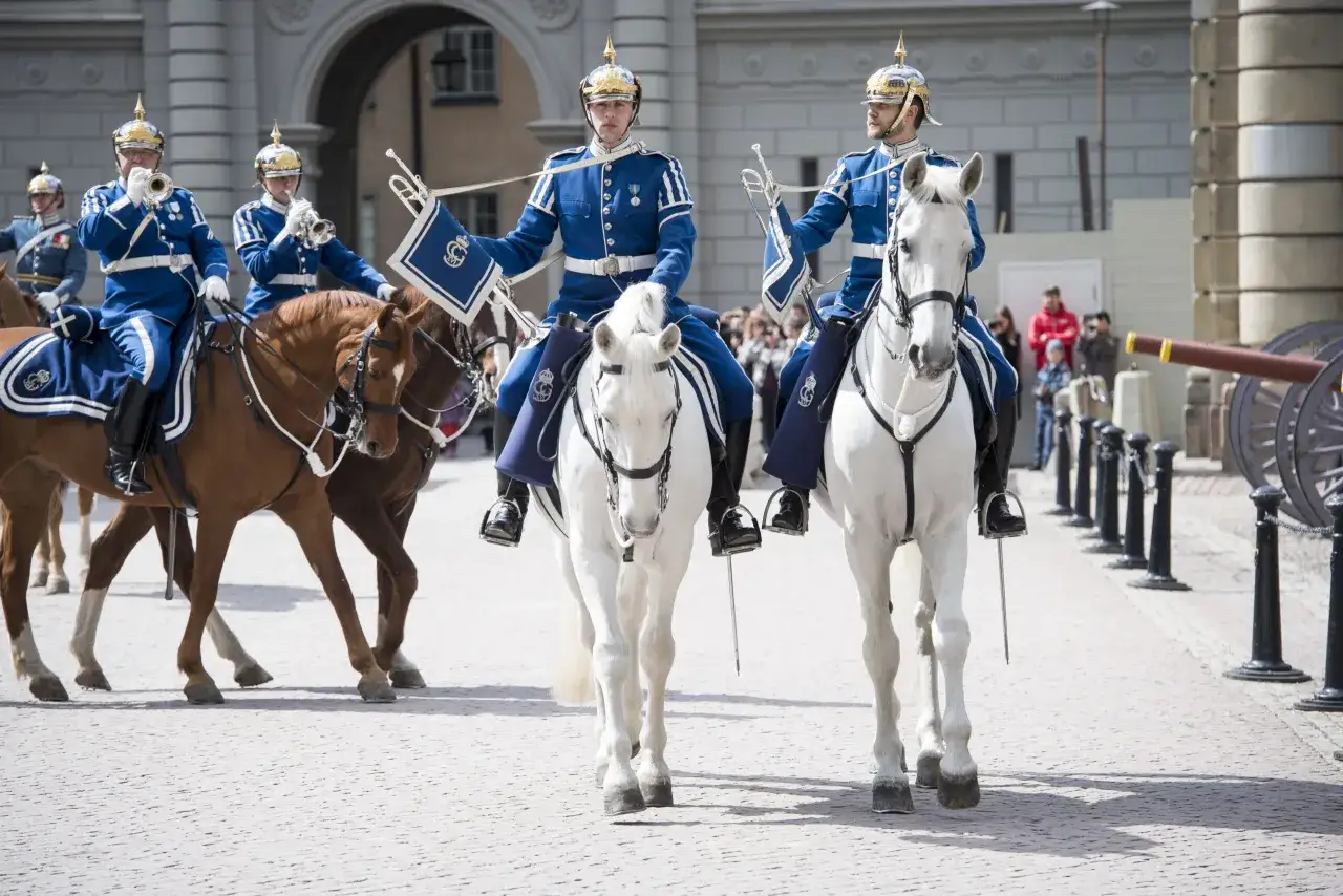 Livgardets dragonmusikk&aring;r i bl&aring; uniformer rider vita och bruna h&auml;star. En musiker spelar trumpet.