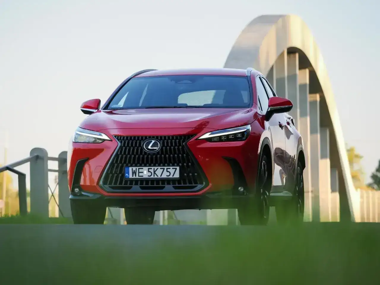 Czerwony suv Lexus NX z charakterystycznym grillem i nowoczesnymi światłami stoi przed łukowatym mostem.