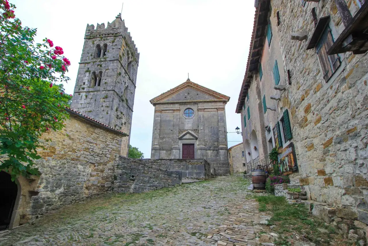 Steile Kopfsteinpflasterstraße führt zu einer Kirche und einem Glockenturm in Hum, Kroatien. Rote Rosen blühen neben alten Steinhäusern.