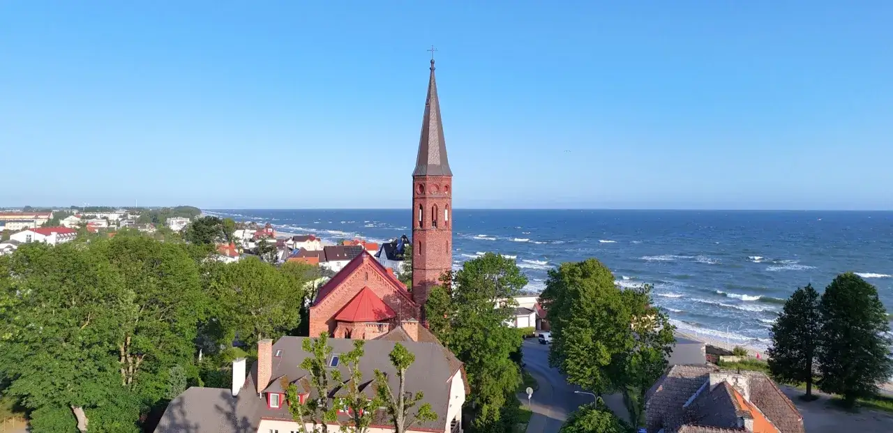 Widok na kości&oacute;ł i morze w Sarbinowo centrum. Dachy dom&oacute;w, zielone drzewa i błękitne niebo.