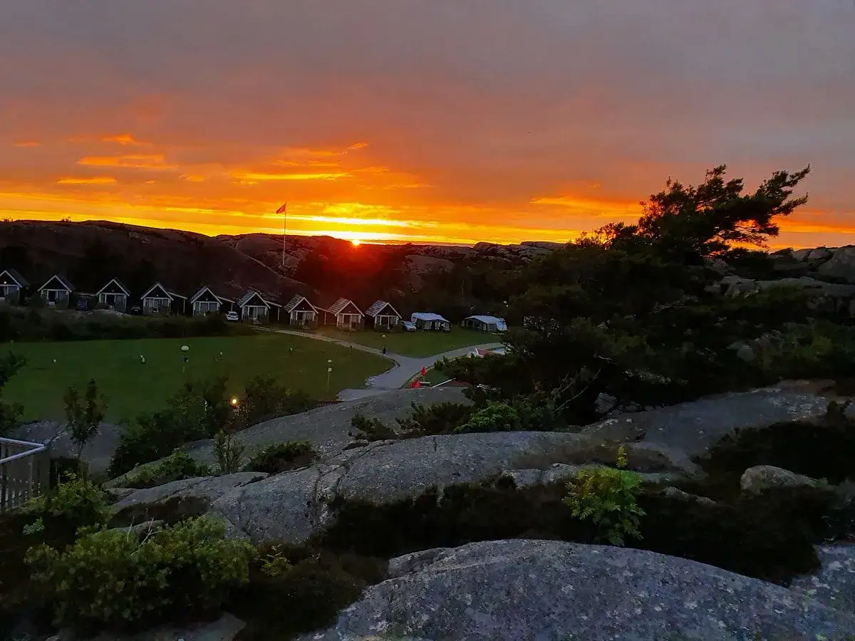 Solvik camping Smögen badar i solnedgångens glöd. Små stugor längs en grönskande gräsyta under en dramatisk himmel.