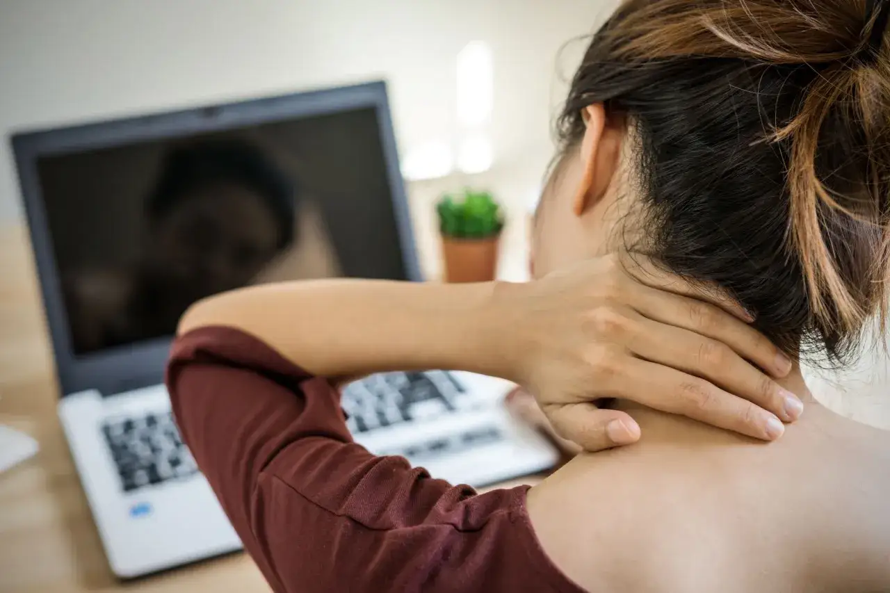 Kobieta masuje kark, odczuwając ból kręgów szyjnych podczas pracy przy laptopie.