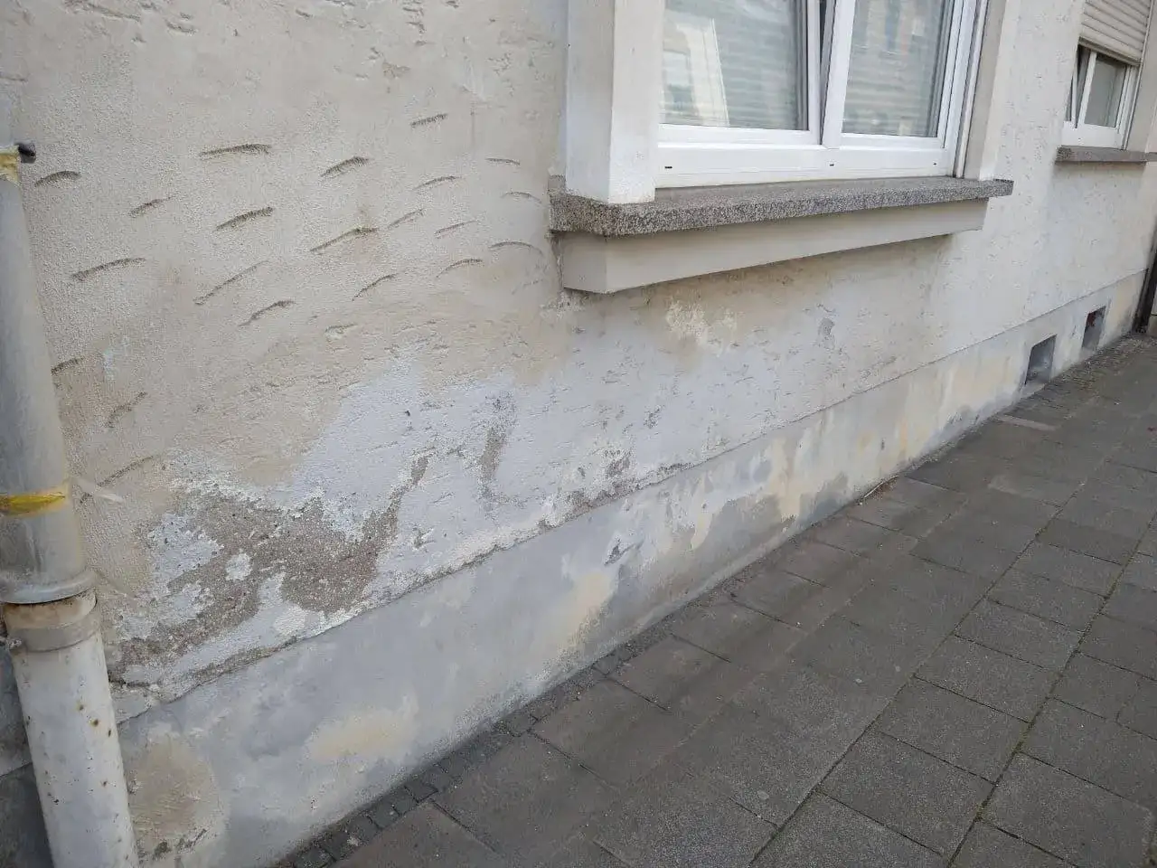 Salpeter-Schäden am Mauerwerk zeigen sich als weiße Ausblühungen und abblätternder Putz an der Hauswand neben dem Fenster.