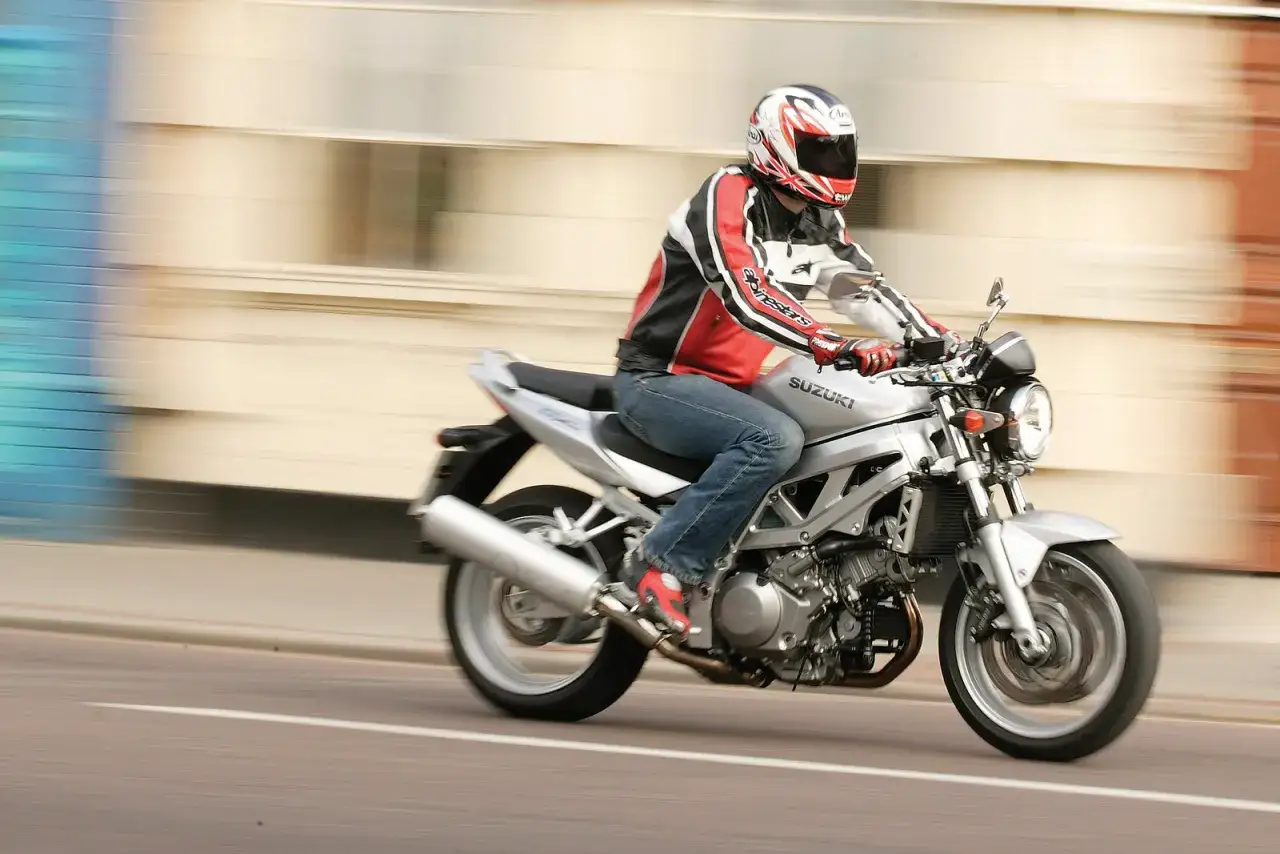 Motocyklista w kasku i skórzanej kurtce pędzi na srebrnym Suzuki SV1000.