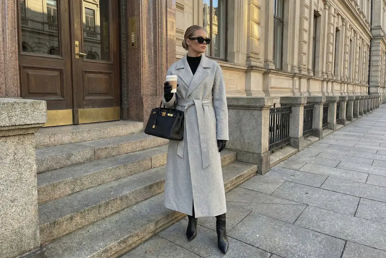 Elegancka kobieta w szarej bosmance, trzymająca kawę i torebkę, stoi na schodach przed budynkiem.