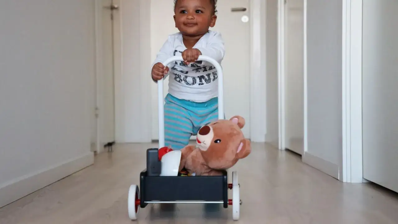 Bambino sorridente impara quando iniziano a camminare i bambini, spingendo un carrellino con un orsacchiotto.