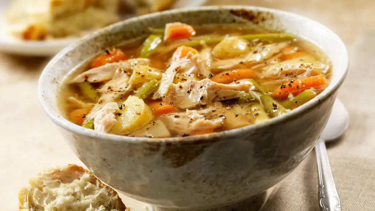 Una ciotola fumante di zuppa di pollo con carote, patate e fagiolini, condita con pepe nero. Accanto, una fetta di pane croccante.