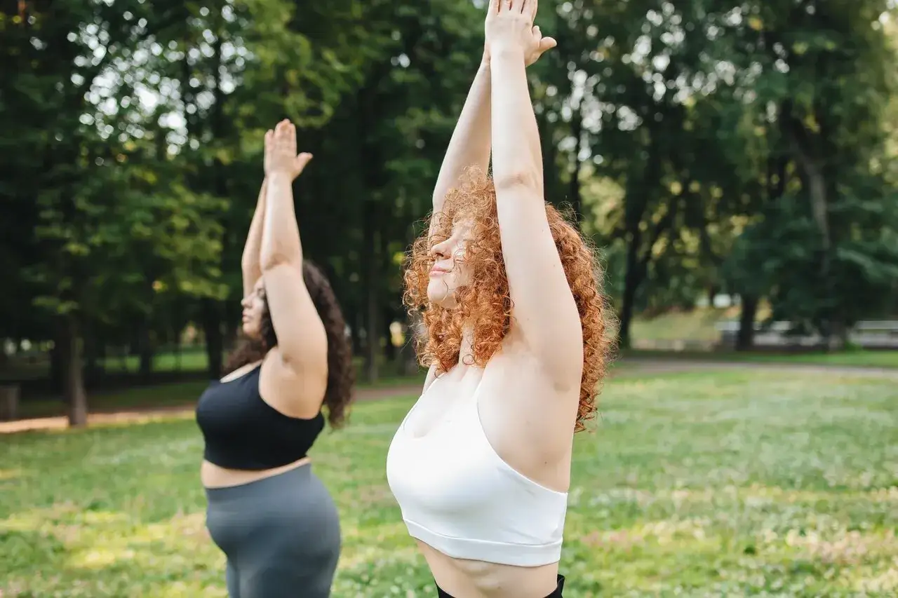 Dos mujeres hacen yoga en un parque. La mujer de la derecha tiene el pelo rizado y rojo. Se puede hacer ejercicio con la regla, sin importar tu talla.