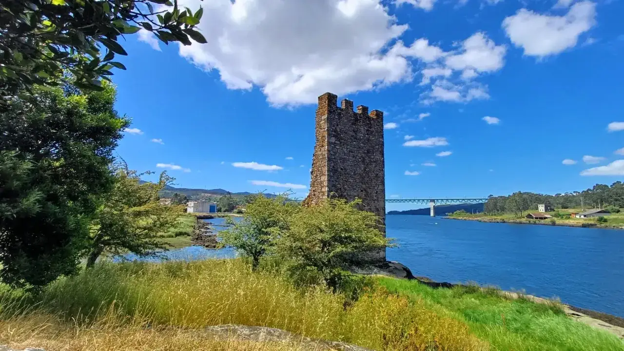 Torre medieval junto al r&iacute;o en Catoira, Galicia. Un puente cruza el agua azul bajo un cielo con nubes blancas.