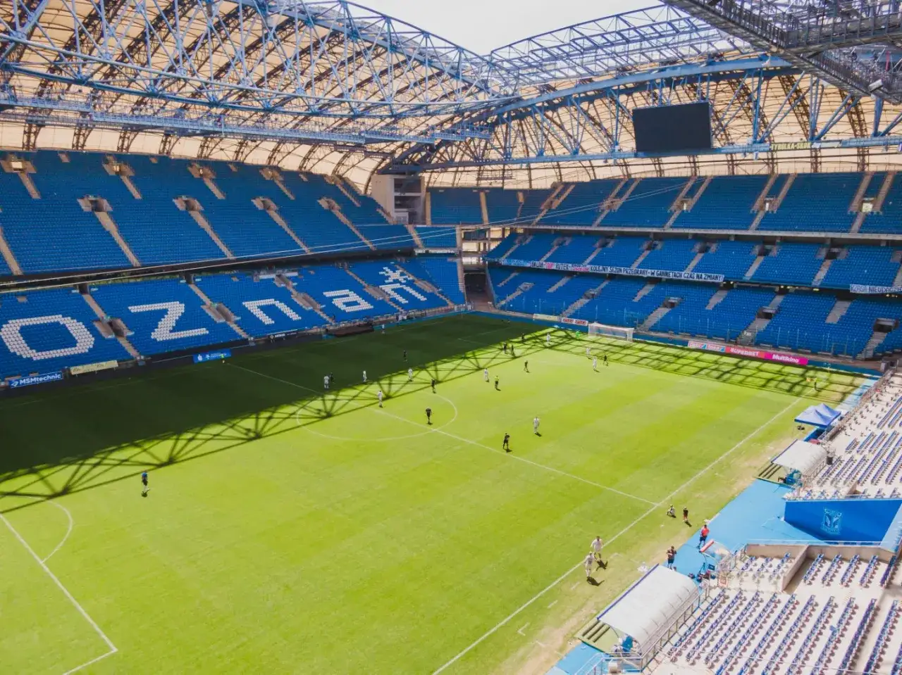 Enea Stadion Poznań z błękitnymi trybunami i zieloną murawą, na kt&oacute;rej trenują piłkarze.