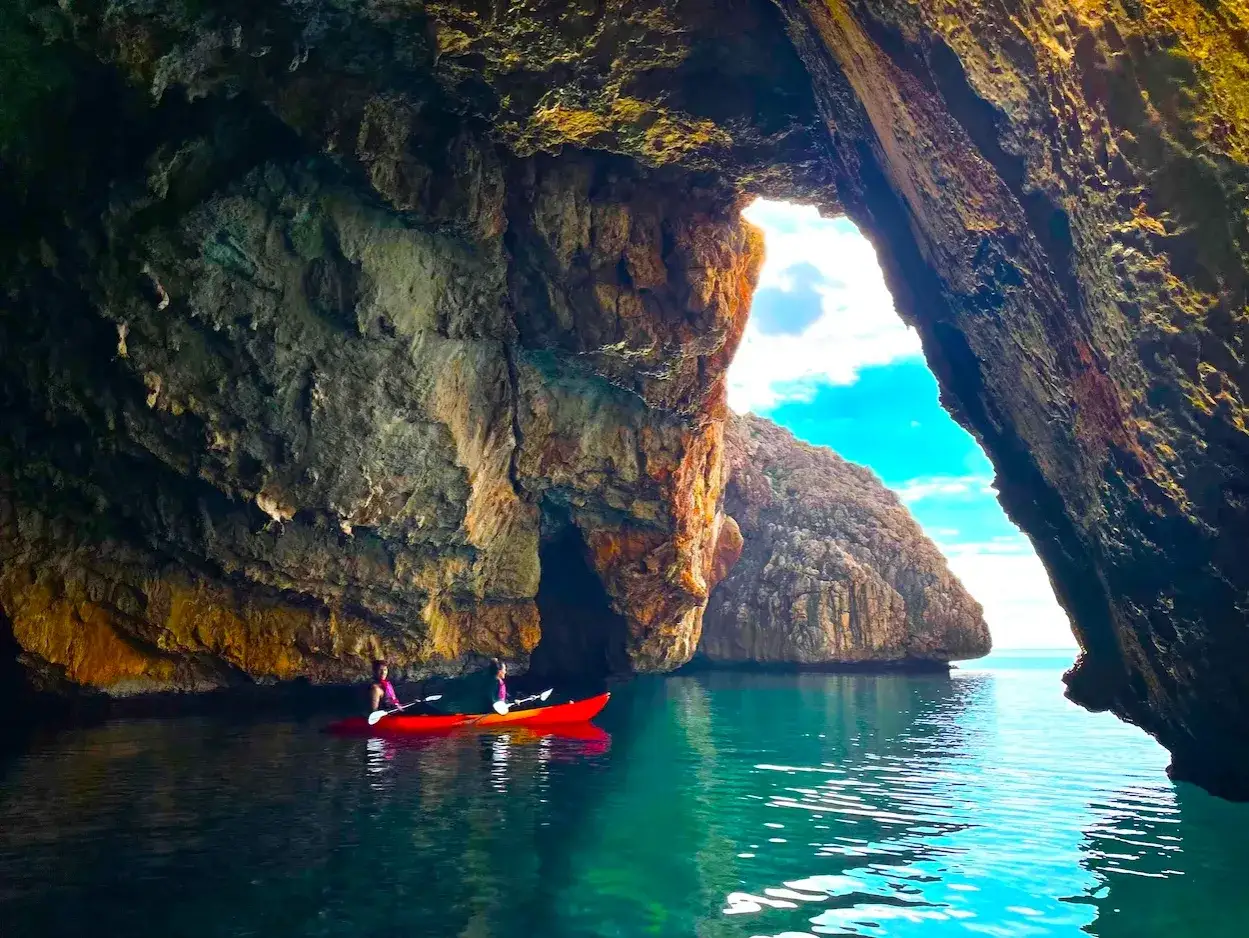 Dos personas en kayak rojo exploran la Cova del Llop Mari en Jávea, con aguas turquesas y formaciones rocosas impresionantes.