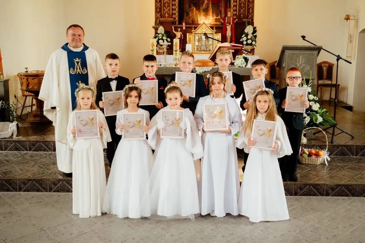 Dzieci w odświętnych strojach, z księdzem, pozują do pamiątkowego zdjęcia po uroczystości komunii świętej.