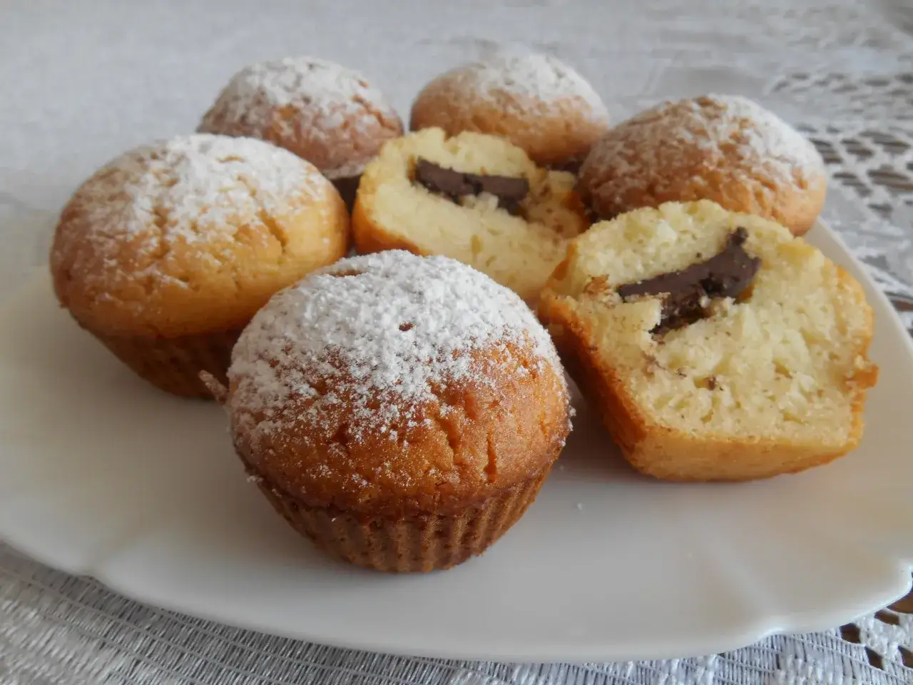 Pyszne muffinki waniliowe z czekoladą, oprószone cukrem pudrem, kuszą swoim wyglądem. Jeden z nich przekrojony, ukazuje kawałek gorzkiej czekolady.