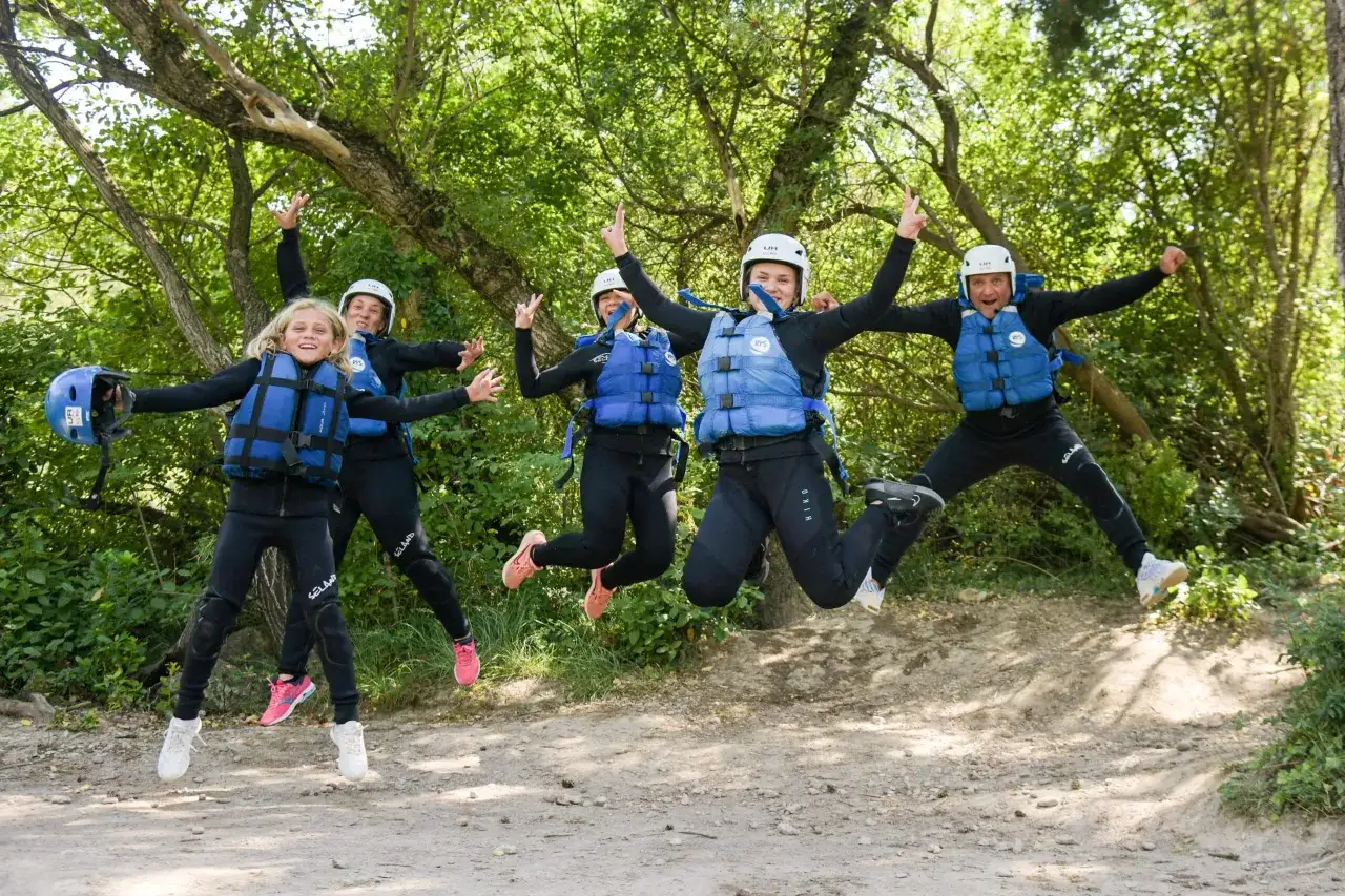 Un grupo de cinco personas, incluyendo ni&ntilde;os, salta de alegr&iacute;a con trajes de neopreno y chalecos salvavidas en un entorno natural, listos para una aventura en Huesca.