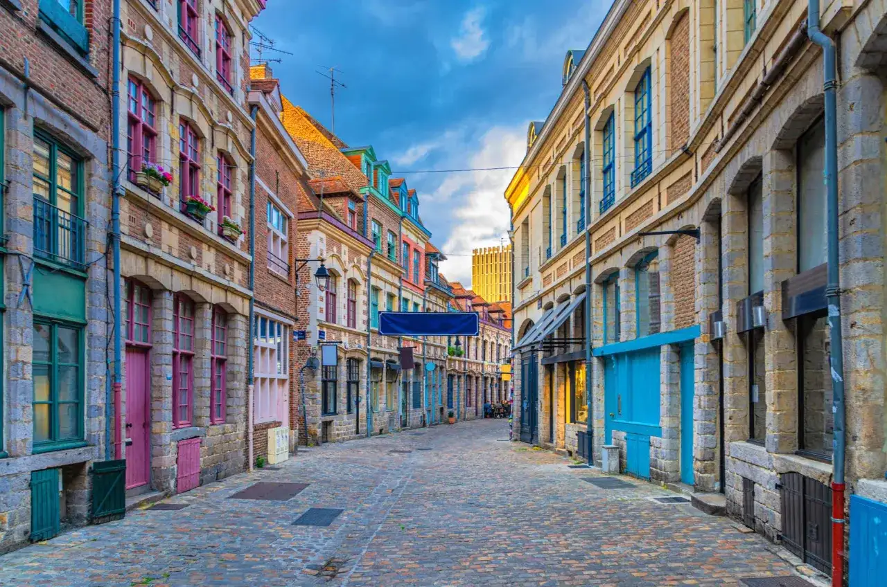 Une ruelle pavée bordée de maisons colorées, typiques des plus belles rues du Vieux-Lille. Le ciel bleu et nuageux contraste avec les façades aux portes et fenêtres vives.