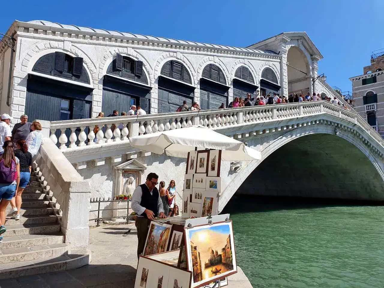 Un artiste expose ses tableaux de Venise, payant, sous le pont du Rialto. L'eau émeraude reflète le ciel bleu.