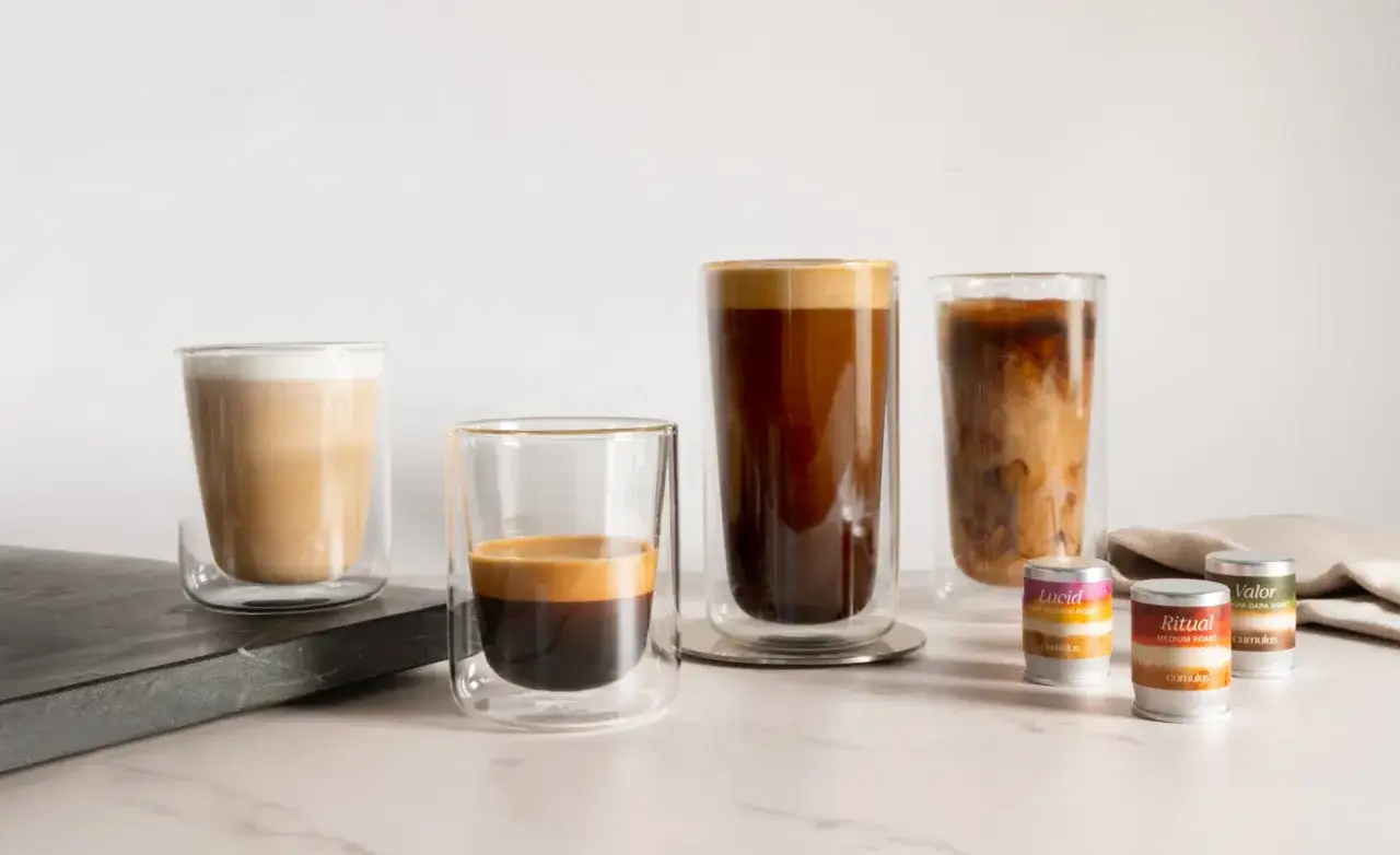 Ein Cortado Kaffee, serviert in einem doppelwandigen Glas, neben anderen Kaffeespezialitäten und Kapseln.