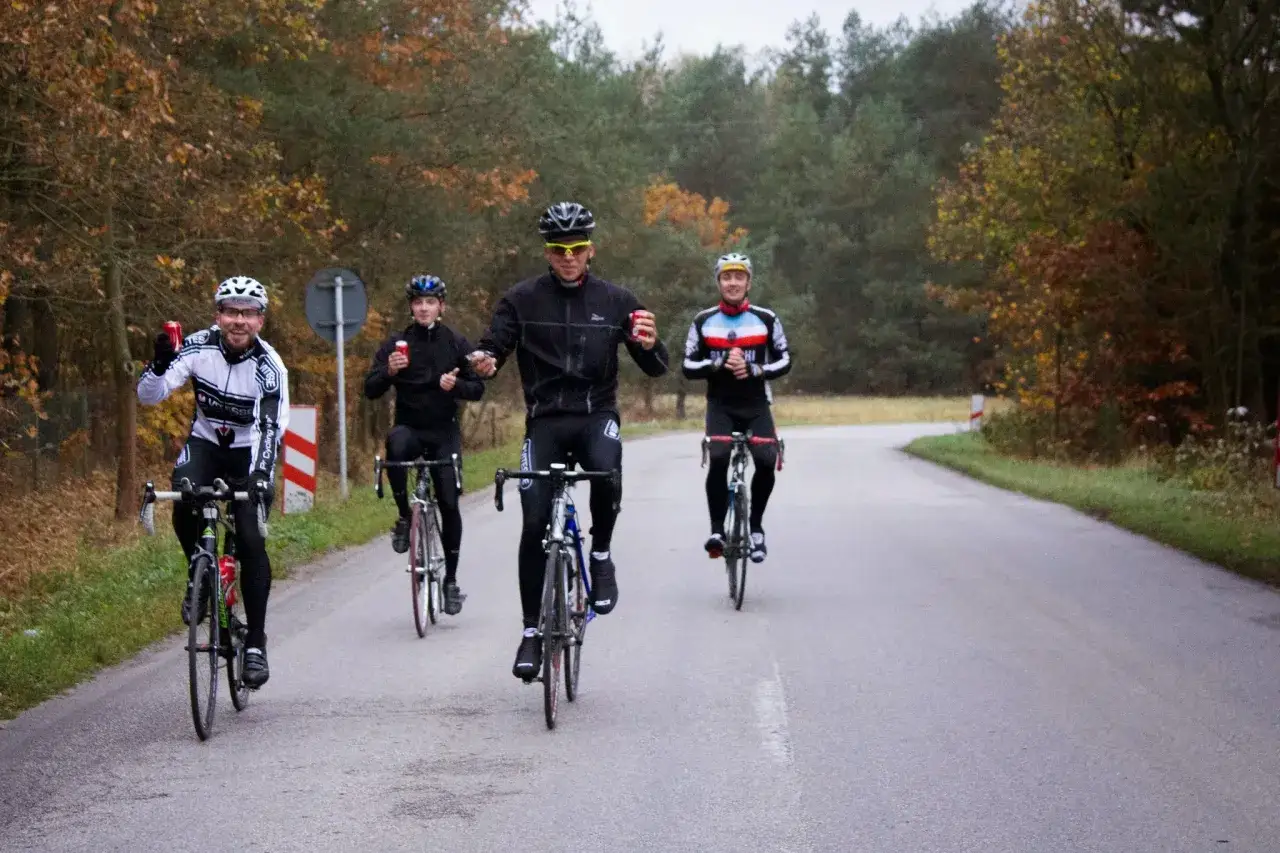 Peleton kolarzy jedzie po jesiennej drodze. Wszyscy w kaskach i strojach kolarskich, jeden z nich macha do aparatu.