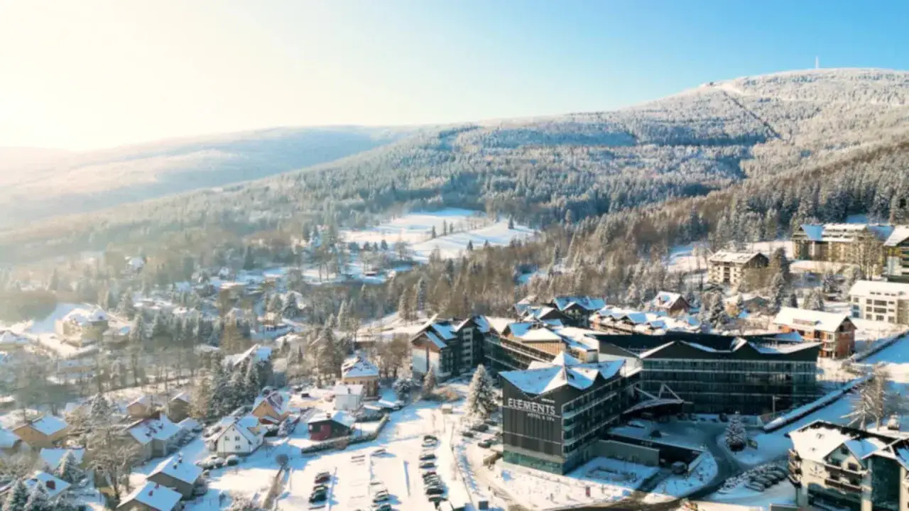 Hotel ze stokiem w zimowej scenerii. Śnieg pokrywa dachy domów i drzewa, tworząc malowniczy krajobraz.