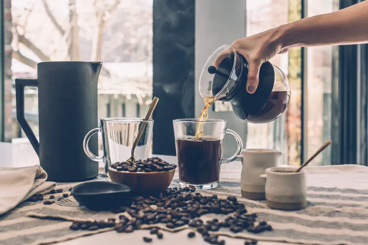 La produzione del caffè in corso: caffè versato da una caraffa in una tazza, chicchi sparsi, brocca scura e tazze in ceramica.
