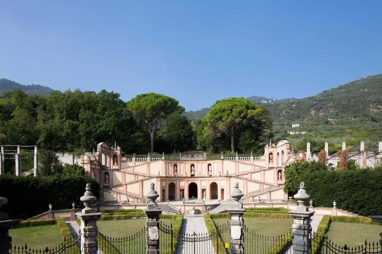 La Villa Bettoni, con le sue imponenti scalinate e statue, si staglia contro un cielo azzurro, circondata da un giardino curato e colline verdi.