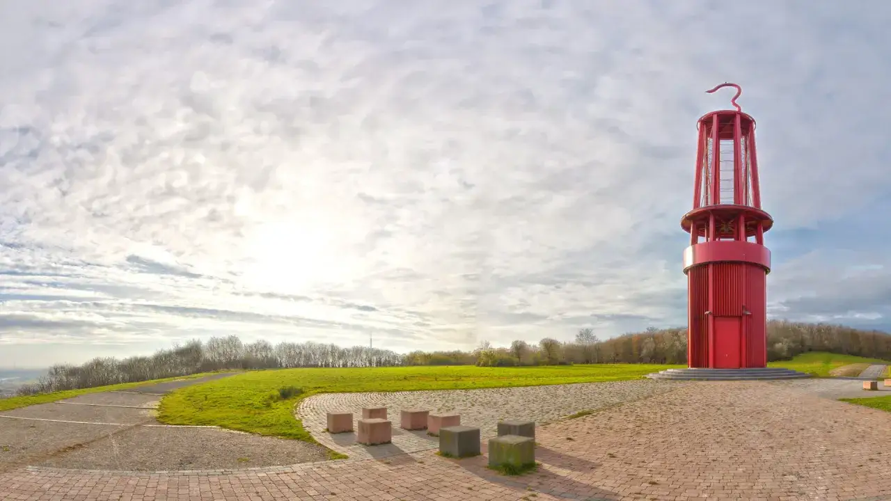 Rote Aussichtsturm auf der Halde Moers, umgeben von gr&uuml;nen H&uuml;geln und einem weiten Himmel mit Wolken.