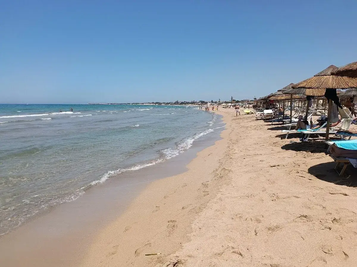Plage de sable fin à **letojanni sicile**, avec des parasols de chaume et des transats. La mer est calme et le ciel est bleu.