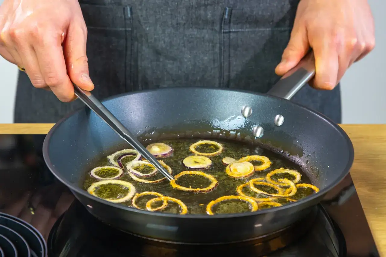 Zwiebelringe in heißem Öl in der Pfanne, um sie knusprig zu braten. Eine Hand mit einer Zange wendet die Ringe.