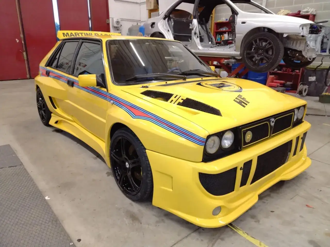 Żółta Lancia Delta Integrale z paskami Martini Racing w warsztacie, obok rozbierany biały samochód.
