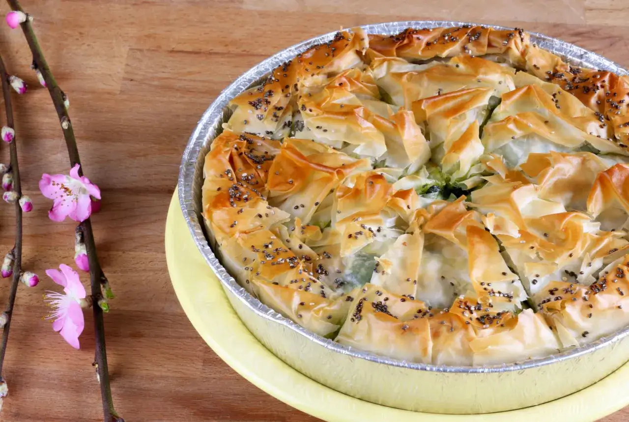 Złocista spanakopita z chrupiącym ciastem filo, posypana makiem, na drewnianym tle obok gałązki z różowymi kwiatami.