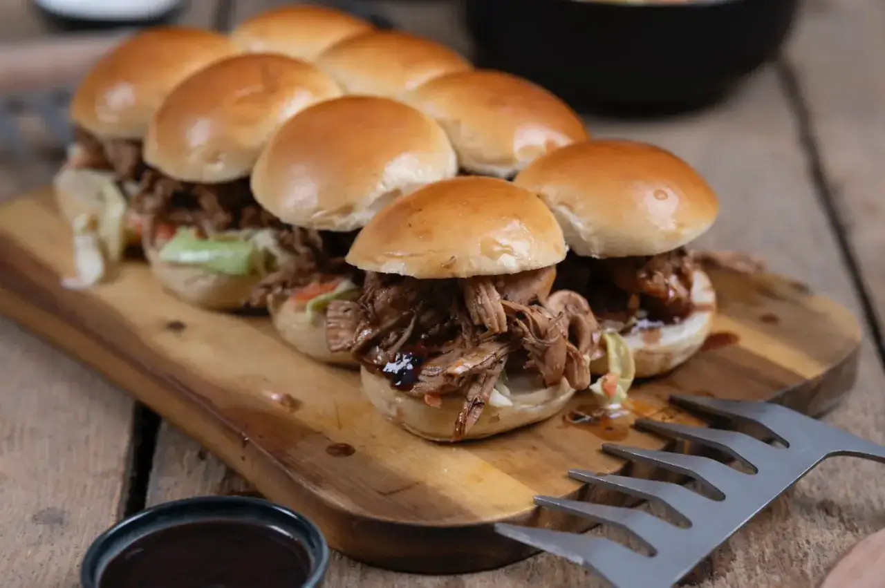 Saftiges pulled chicken im Backofen zubereitet, serviert in kleinen Brötchen auf einem Holzbrett.