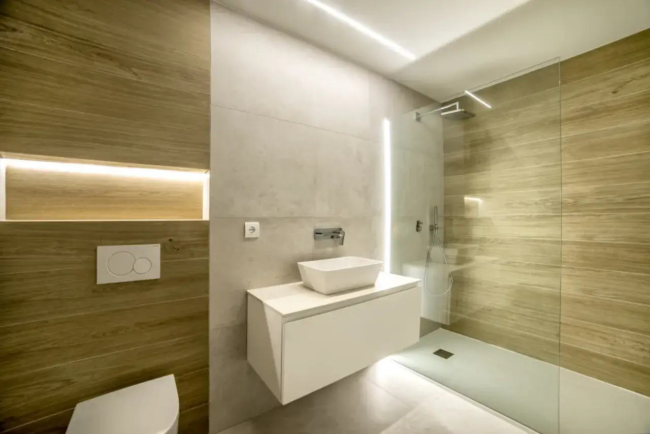 Baño moderno con revestimiento de madera y **pladur para baños**. Lavabo sobre mueble flotante, ducha de cristal y WC empotrado.