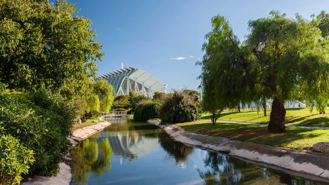 Un canal serpentea entre vegetación exuberante, reflejando el cielo azul y la futurista arquitectura de la Ciudad de las Artes y las Ciencias, uno de los principales lugares turísticos de Valencia.